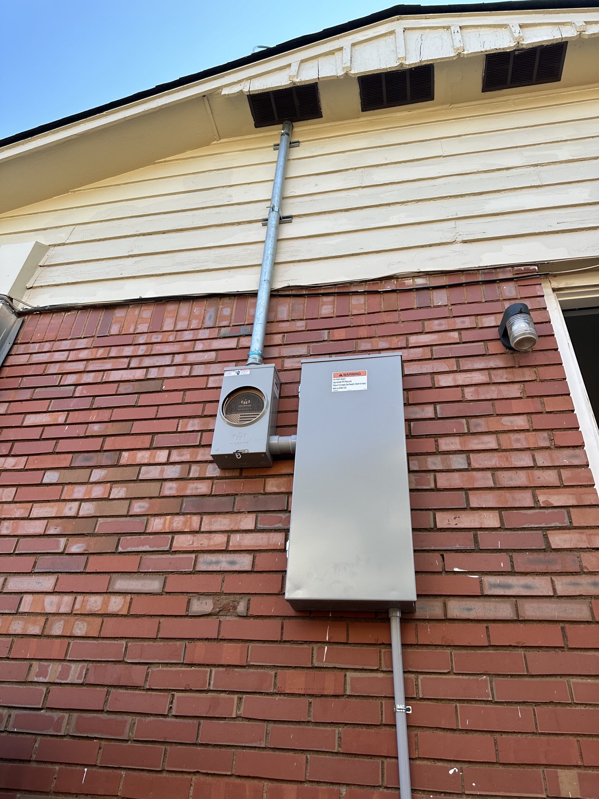A brick wall with an electric meter, breaker box, and conduit pipes attached sits below a light fixture on the exterior of a house—ideal for electrical panel upgrades and repairs.