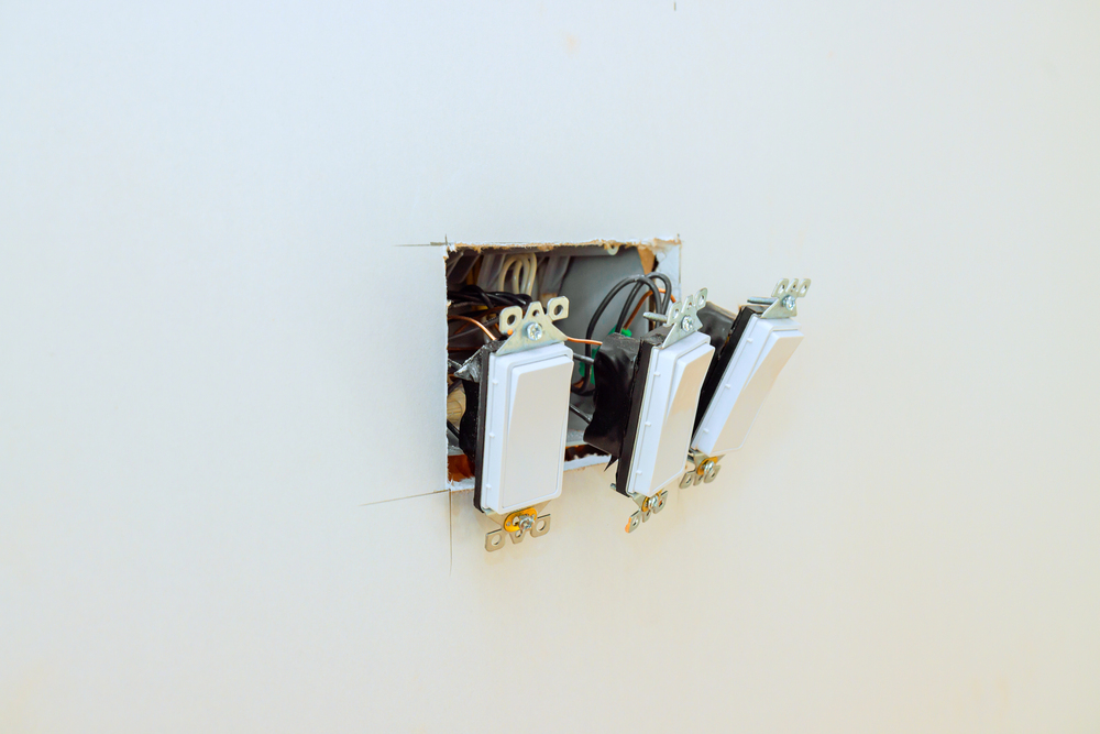 Three light switches partially installed in an open wall box with exposed wires, against a plain white wall.